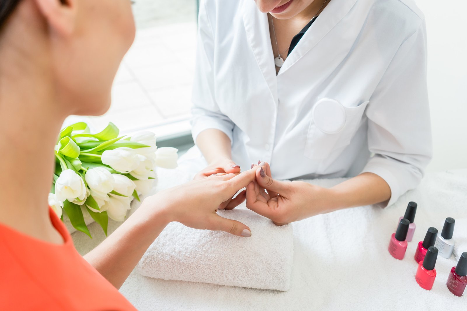 Experienced nail technician wearing white coat while checking the trendy manicure of a young client in a modern beauty salon