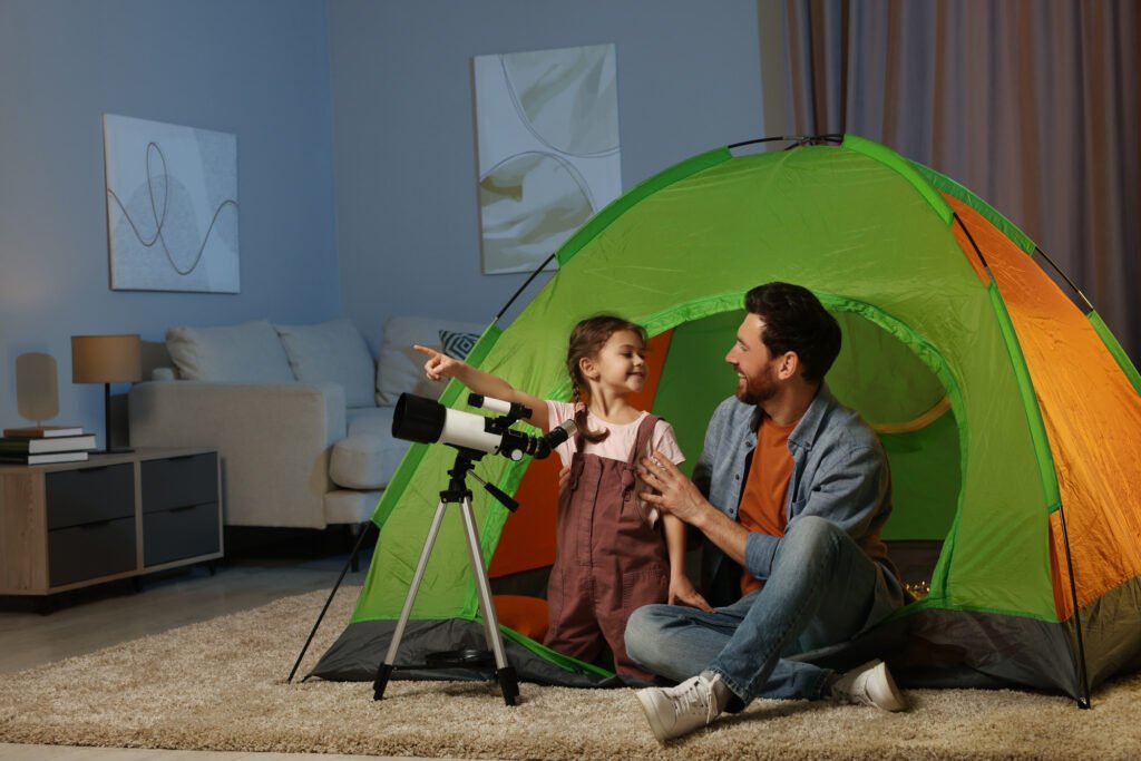 Father and his daughter using telescope to look at stars while sitting in camping tent indoors