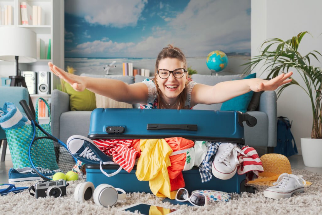 Happy woman leaning on her overpacked trolley suitcase with outstretched arms and smiling at camera, she is getting ready for a trip