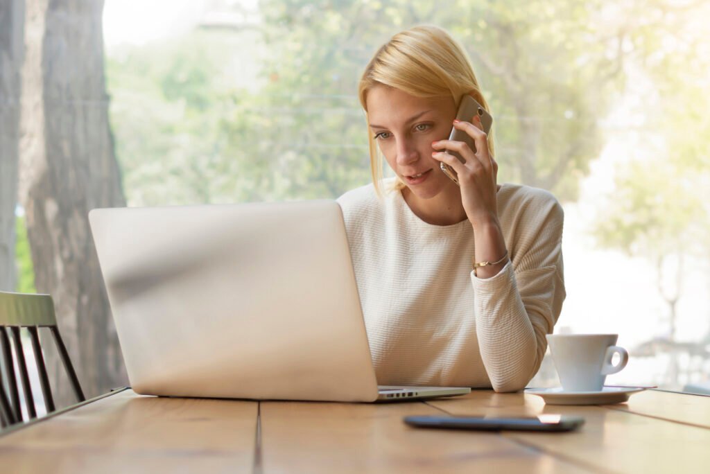 Portrait of experienced young businesswoman at work break keyboarding text on laptop computer while talking on smartphone, female person busy working in modern office interior or coffee shop, flare