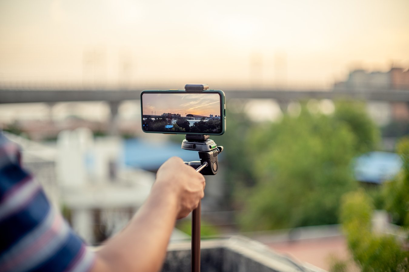 mobile phone mounted on a tripod at full extention replacing a DSLR for a professional shoot to capture a sunset with a metro station and train arriving as seen on the screen with rest out of focus. Shows the rapid advancement of technology that allows people to use their mobile phones for professional things like vlogging, timelapse, hyperlapse, youtube and tiktok videos