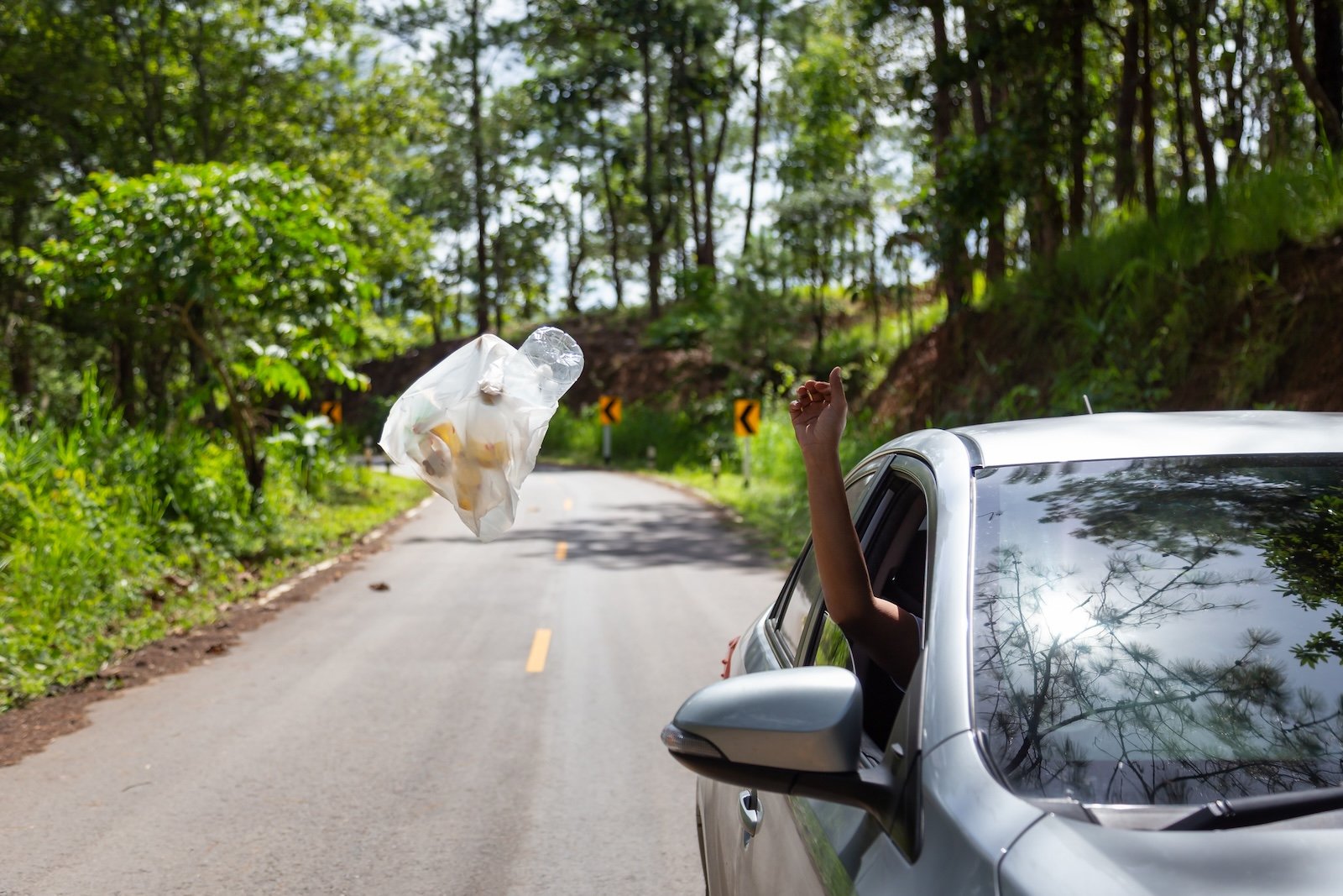 Male driver throwing trash from car window on the road,man’s hand or arm throwing garbage bag out of car window to the floor while driving on valley road in green nature,pollute with plastic waste,environmental protection