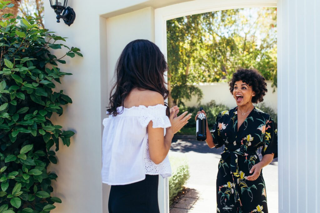 Excited african woman attending a female friend's housewarming party. Woman giving a wine bottle her friends at entrance door.