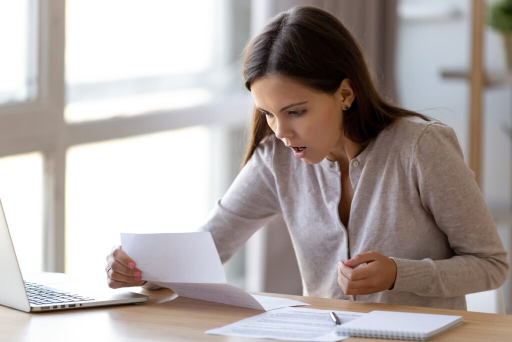 Surprised,Young,Woman,Sitting,At,Desk,Holding,Paper,Reading,Unbelievable
